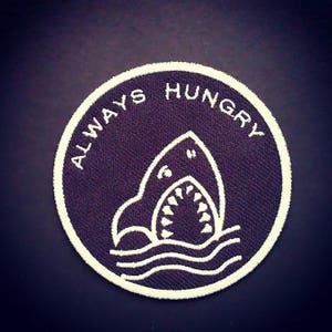 Puede incluir: Un parche circular con fondo azul marino y detalles bordados en blanco. El parche presenta un tiburón con la boca abierta sobre líneas onduladas, y las palabras "ALWAYS HUNGRY" están bordadas alrededor del borde.