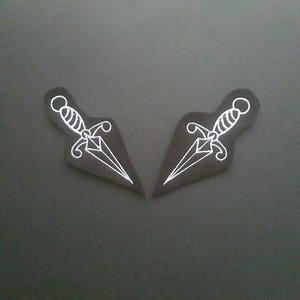 Può includere: Due toppe da stirare ricamate bianche a forma di pugnali su uno sfondo nero.