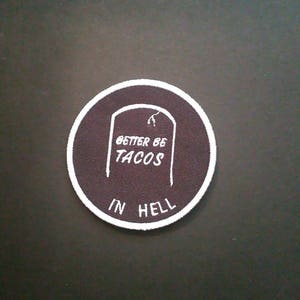 Può includere: Un cerotto rotondo con bordo bianco e centro marrone. Il cerotto presenta una lapide bianca con il testo "Better Be Tacos" e le parole "In Hell" sotto la lapide.