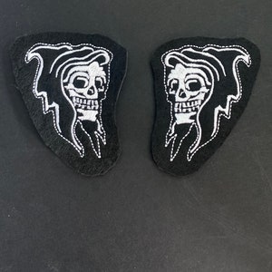May include: Two black embroidered patches featuring a white skull design with a flowing hood and lightning bolt accents. The patches are shaped with a curved edge and are suitable for sewing or ironing onto fabric.