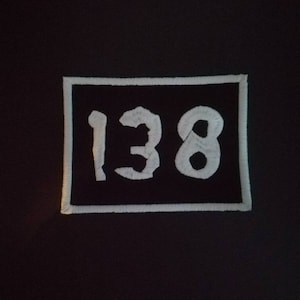 May include: A black embroidered patch featuring the number "138" in white. The numbers are in a bold, slightly distressed font, and are centered within a white-bordered black rectangle. The patch is likely for clothing or accessories.