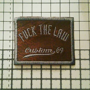 May include: A brown leather patch with a gray embroidered border. The patch says "F**K THE LAW" in large letters and "Custom 69" in smaller letters.