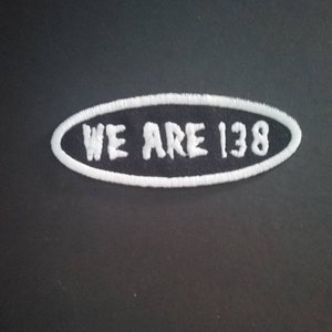 Może przedstawiać: Biała naszywka haftowana z czarnym owalnym tłem. Tekst "WE ARE 138" jest wyszyty na biało.