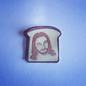 Könnte beinhalten: Ein gestickter Aufnäher mit einem Gesicht auf einem weißen Brot Hintergrund.