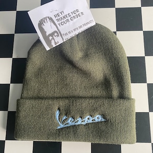 Puede incluir: Un gorro de punto verde oliva con el logotipo "Vespa" bordado en azul claro. Una pequeña tarjeta blanca con el texto "HEY! THANKS FOR YOUR ORDER" está metida en el gorro. El gorro se muestra sobre una superficie a cuadros blancos y negros.