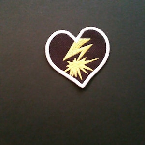 Peut inclure: Écusson en forme de cœur avec un fond noir, une bordure blanche et un motif d'éclair et d'éclat jaune. Idéal pour les vêtements ou les accessoires.