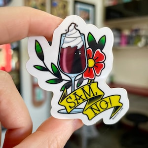 May include: A white sticker with a red flower and green leaves. A glass of red wine with whipped cream on top is in the center of the sticker. A yellow banner with the text "SAM, NO!" is below the glass.