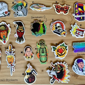 Puede incluir: Una colección de pegatinas pequeñas con varios diseños, incluyendo una lápida con la palabra "ANXIETY" en ella, un cuervo con ojos rojos, una calavera con llamas, una mariposa con alas rojas, un esqueleto con una patineta y un lobo con llamas. Las pegatinas están sobre una superficie de madera.