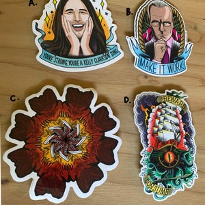 Könnte beinhalten: Eine Sammlung von vier großen Aufklebern mit verschiedenen Designs. Einer zeigt eine lächelnde Person mit dem Text "YOU'RE STRONG, YOU'RE A KELLY CLARKSON SONG." Ein anderer zeigt einen Mann mit dem Text "I MAKE IT WORK." Weitere Aufkleber zeigen ein Blumenmuster und ein nautisches Tattoo.