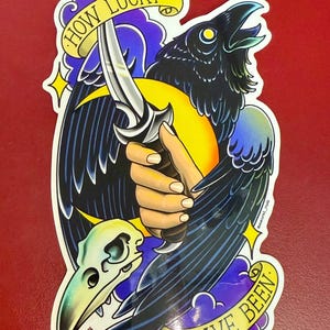 May include: A black and blue crow with a yellow crescent moon behind it. A hand holds a silver dagger with a black handle. A white skull with a blue and green beak is below the crow. The text "HOW LUCKY I HAVE BEEN." is in a yellow banner.