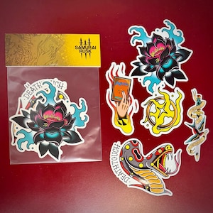 May include: A collection of tattoo-style stickers featuring a black lotus flower with pink and teal accents, a snake, a hand holding a card, a starburst, and a dagger. The stickers are on a red background. The brand name "SAMURAI RUSK" is visible.