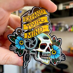 May include: A sticker with a skull and a wrench, with the words "Expand Your Mind" on a yellow banner. The skull is blue and white, and the wrench is black and silver. The sticker is surrounded by flowers.
