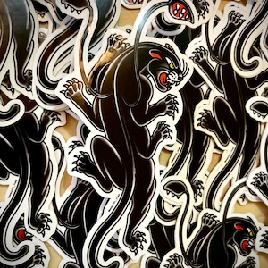 May include: A close-up of a stack of black panther stickers. The stickers are printed on white paper and feature a black panther with a white outline. The panther is in a crouching position with its claws extended.