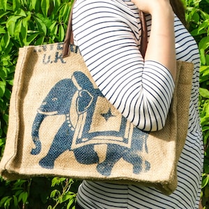 Könnte beinhalten: Eine braune Jute-Tragetasche mit einem blauen Elefanten-Design. Die Tasche hat einen braunen Ledergriff und den Text "U.K." auf der Vorderseite.