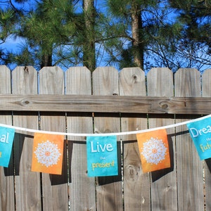 May include: A decorative banner with orange and teal flags strung on a white cord. Each flag has a white mandala design. The teal flags have the phrases "Heal the past", "Live the present", and "Dream the future". The banner is hanging on a wooden fence.