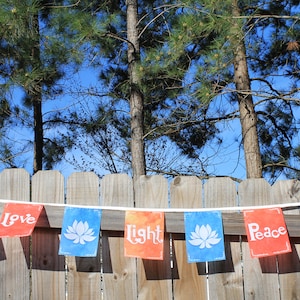 May include: A banner with rectangular flags strung across a wooden fence. The flags are blue, orange, and red, with the words "Love", "Light", and "Peace" printed on them. Each flag has a white lotus flower design.