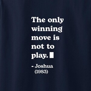Può includere: T-shirt blu navy con la scritta bianca: "The only winning move is not to play." attribuita a Joshua (1983). La maglietta ha girocollo e maniche corte.