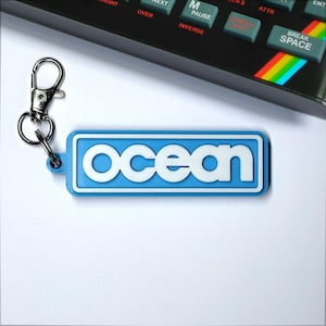 Puede incluir: Un llavero rectangular azul con la palabra "ocean" en letras blancas. Cuenta con un mosquetón y un anillo de metal plateado. Un dispositivo electrónico negro con una franja de arcoíris está en el fondo.