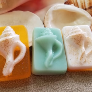May include: Three bars of soap shaped like seashells. The soap on the left is orange with a white seashell, the middle soap is light blue with a white seashell, and the soap on the right is white with a white seashell.