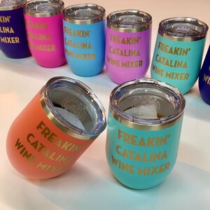 May include: A set of 10 colorful stainless steel wine tumblers with a gold inscription that reads "Freakin' Catalina Wine Mixer". The tumblers are in various colors, including blue, pink, teal, orange, and purple. The tumblers are all the same size and shape, and they have a lid with a straw hole. The tumblers are arranged in a semi-circle on a white surface. The text "*Hand Wash Only*" is printed below the tumblers.