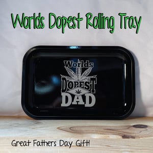 May include: A black rolling tray with the text "Worlds Dopest Dad" in silver. The tray has a rectangular shape with rounded corners. The words "Worlds Dopest Rolling Tray" and "Great Fathers Day Gift!" are also visible.