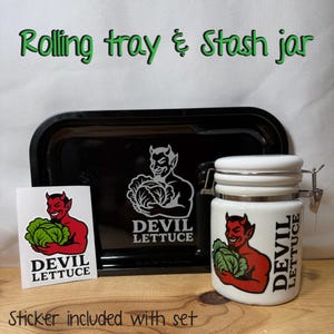 May include: A black rolling tray and stash jar set. The set includes a black rectangular tray, a white jar with a metal clasp, and a sticker. Each item features a devil graphic holding lettuce and the text "DEVIL LETTUCE".