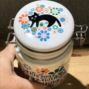 May include: White ceramic jar with a black cat and colorful flowers on the lid. The jar has a metal latch and the text "This may or may not be a nip" on the side.