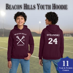 May include: Maroon youth hoodie with the text "BEACON HILLS" above a lacrosse stick graphic. The back of the hoodie displays "STILINSKI" and the number "24". The hoodie is paired with blue jeans. The image also includes the text "NAME & NUMBER COMBOS".