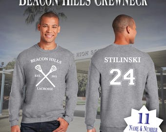 May include: Gray crewneck sweatshirt with white text and graphics. The front displays "BEACON HILLS LACROSSE" with crossed lacrosse sticks and "EST. 2011." The back shows "STILINSKI 24." The image also shows "11 NAME & NUMBER CHOICES."