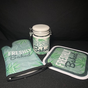 May include: A kitchen set featuring a white ceramic jar with a metal clasp, an oven mitt, and a pot holder. All items have a green cannabis leaf pattern and the words "FRESHLY Baked" in white.