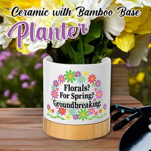May include: White ceramic planter with a bamboo base. The planter has a floral design with the text "Florals? For Spring? Groundbreaking."