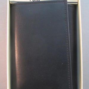 May include: A black leather three-fold wallet by Buxton, presented in a white box. The wallet is closed, showing a smooth, dark surface with stitched edges. The box has the text "Three Fold" and "BUXTON" printed on it, along with "Since 1898".