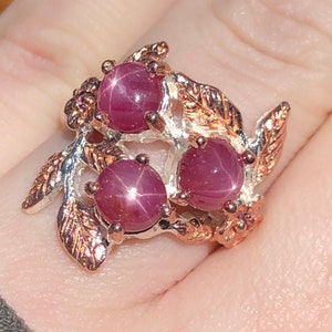May include: A ring featuring three star ruby gemstones set in a silver and rose gold floral design. The gemstones are a deep red color with a star-like effect. The ring has a detailed, nature-inspired aesthetic.