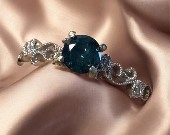Könnte beinhalten: Ein silberfarbener Ring mit einem großen, runden, dunkelblau-türkisfarbenen Mittelstein. Die Ringschiene ist mit komplizierten, geschwungenen Metallmustern verziert, die sich auf beiden Seiten der zentralen Fassung erstrecken und ein detailliertes Aussehen verleihen.