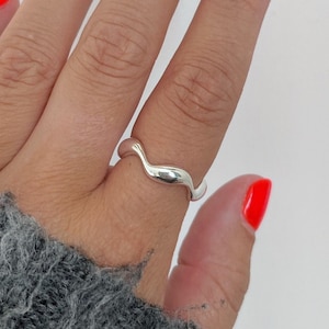 May include: A silver ring with a wavy design is worn on a finger. The ring is smooth and polished. The person's nails are painted a bright orange colour. The person is wearing a grey jumper.