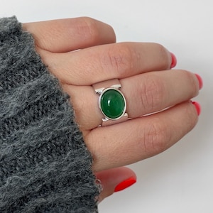 May include: A silver ring featuring a large, oval, vivid green gemstone. The ring has a broad band with a rectangular design either side of the stone. The ring is displayed on a hand with red nail varnish, against a white backdrop.