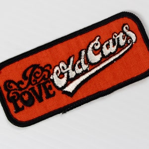 Könnte beinhalten: Ein orangefarbener gestickter Aufnäher mit schwarzen Nähten und weißen Buchstaben, auf dem "I Just Love Old Cars" steht.