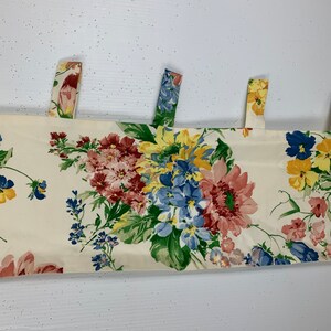 May include: Floral valance with a cream background and colorful flowers in shades of red, blue, yellow, and pink. The valance has three fabric tabs at the top for hanging. The floral pattern includes various types of flowers and green leaves.
