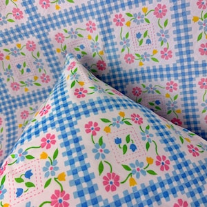 May include: Close-up of a fabric with a blue and white gingham pattern. The fabric is decorated with pink, blue, and yellow floral designs. The flowers are arranged in a repeating pattern, creating a cheerful and inviting aesthetic.