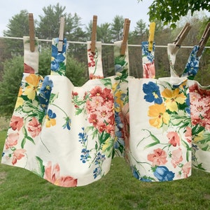 May include: Several floral fabric panels with a cream background are hanging on a clothesline with wooden clothespins. The panels feature a colorful pattern of red, pink, yellow, and blue flowers and green leaves.