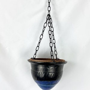 May include: A hanging ceramic planter with a black and blue glazed finish. The planter is suspended by a black chain and features a textured design on the upper portion. The bottom of the planter has a pointed shape.
