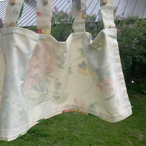 May include: Cream-colored tote bag with floral pattern, hanging from wooden clothespins. The bag features a repeating pattern of pink, yellow, and blue flowers on a cream background. The straps are made of the same floral fabric.