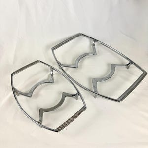 May include: Two chrome-plated metal trivets. Each trivet has a rectangular shape with curved supports and a textured handle. The trivets are designed to protect surfaces from hot cookware.