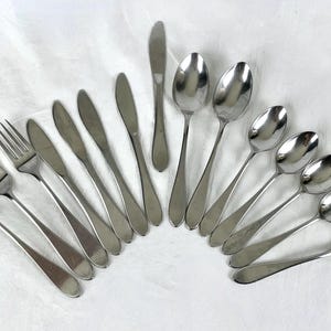 May include: A collection of stainless steel silverware, including forks, knives, and spoons, arranged in a fan shape. The silverware has a polished, reflective surface, and the arrangement suggests a formal dining setting.