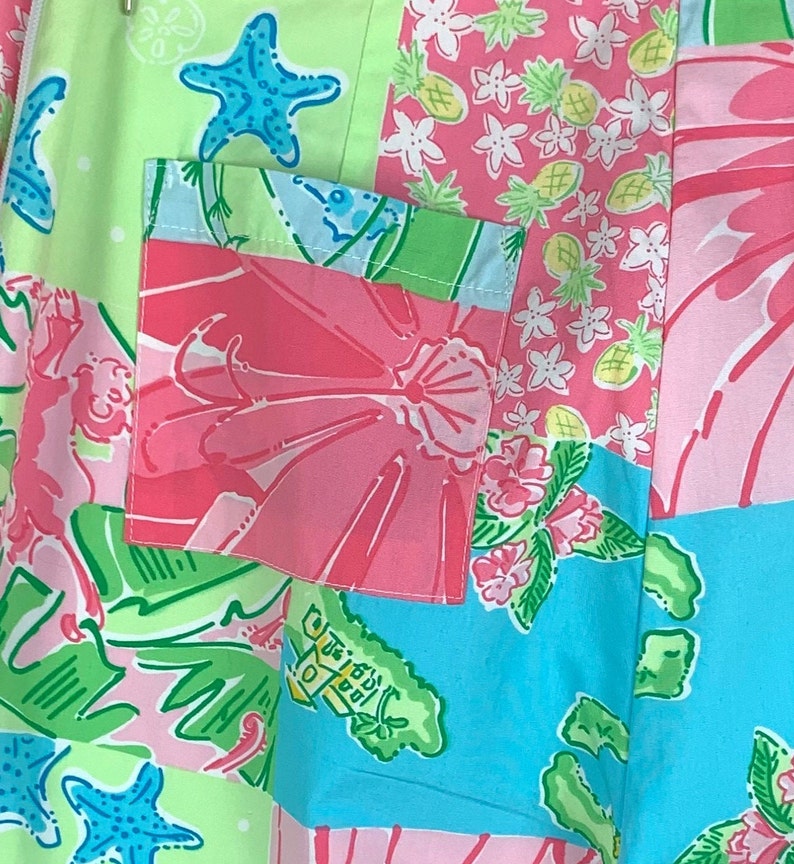 Pu&ograve; includere: Un tessuto patchwork con una variet&agrave; di motivi colorati, tra cui fiori rosa, foglie verdi, stelle marine blu e ananas gialli. Il tessuto ha una tasca blu chiaro con un motivo di fiori rosa.