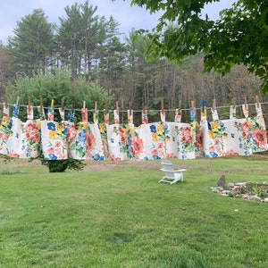 May include: A clothesline displays several floral-patterned aprons, each secured with wooden clothespins. The aprons feature a white background with large, colorful flowers in shades of red, blue, and yellow. The scene is set outdoors with a green lawn and trees.