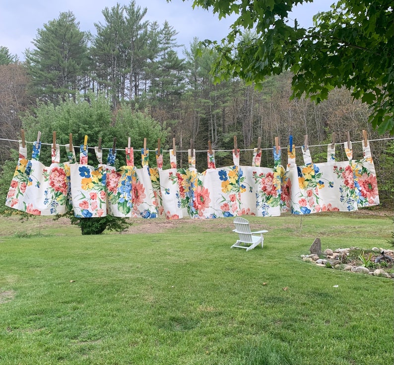 May include: A clothesline displays several floral-patterned aprons, each secured with wooden clothespins. The aprons feature a white background with large, colorful flowers in shades of red, blue, and yellow. The scene is set outdoors with a green lawn and trees.
