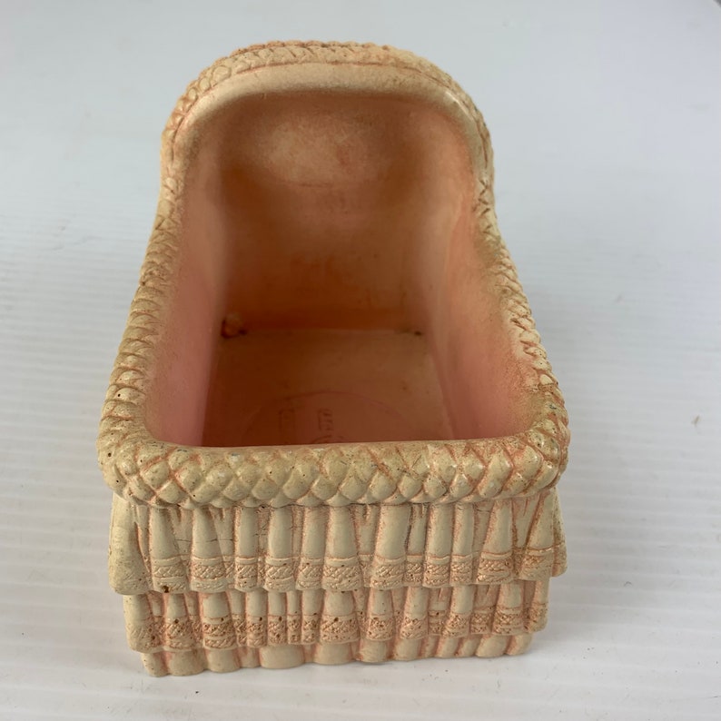 May include: A small, pink ceramic baby crib with a woven design. The crib is empty and ready for a doll or other small toy.
