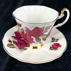 May include: A white teacup and saucer set with a gold trim and a red rose floral pattern.