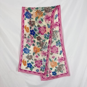 May include: A rectangular scarf featuring a cream-colored background with a vibrant floral pattern in shades of pink, blue, orange, and brown. The scarf is framed by a distinct pink border with a zigzag edge pattern and finished with fringe.